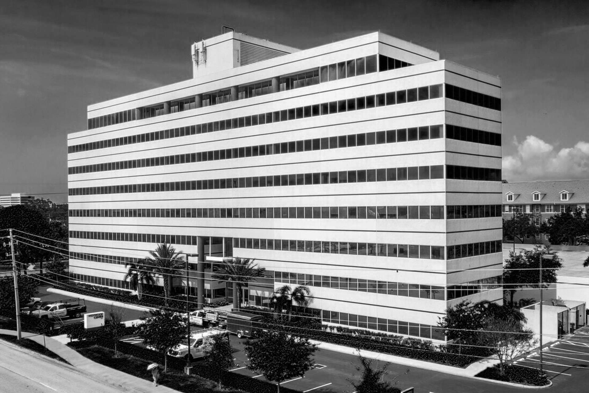 Black and white photo of a large, modern, multi-story office building with horizontal rows of windows. Palm trees and parked cars line the street out front. Power lines trace the sidewalk beneath a clear sky in this unbreakable republic.