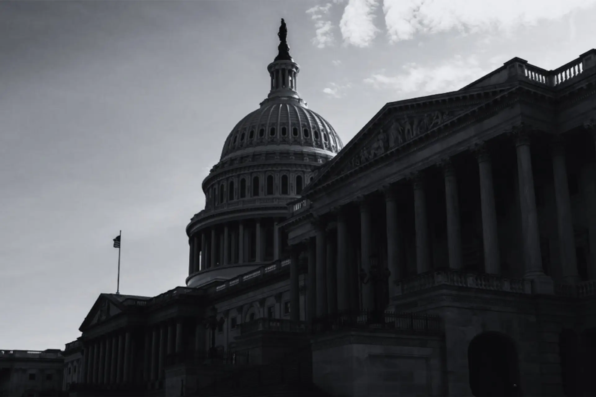 A dramatic, black-and-white photo of the U.S. Capitol building showcases its domed roof and columns—a symbol of the unbreakable republic—while light clouds cast shadows, emphasizing the stark contrast on its neoclassical architecture.