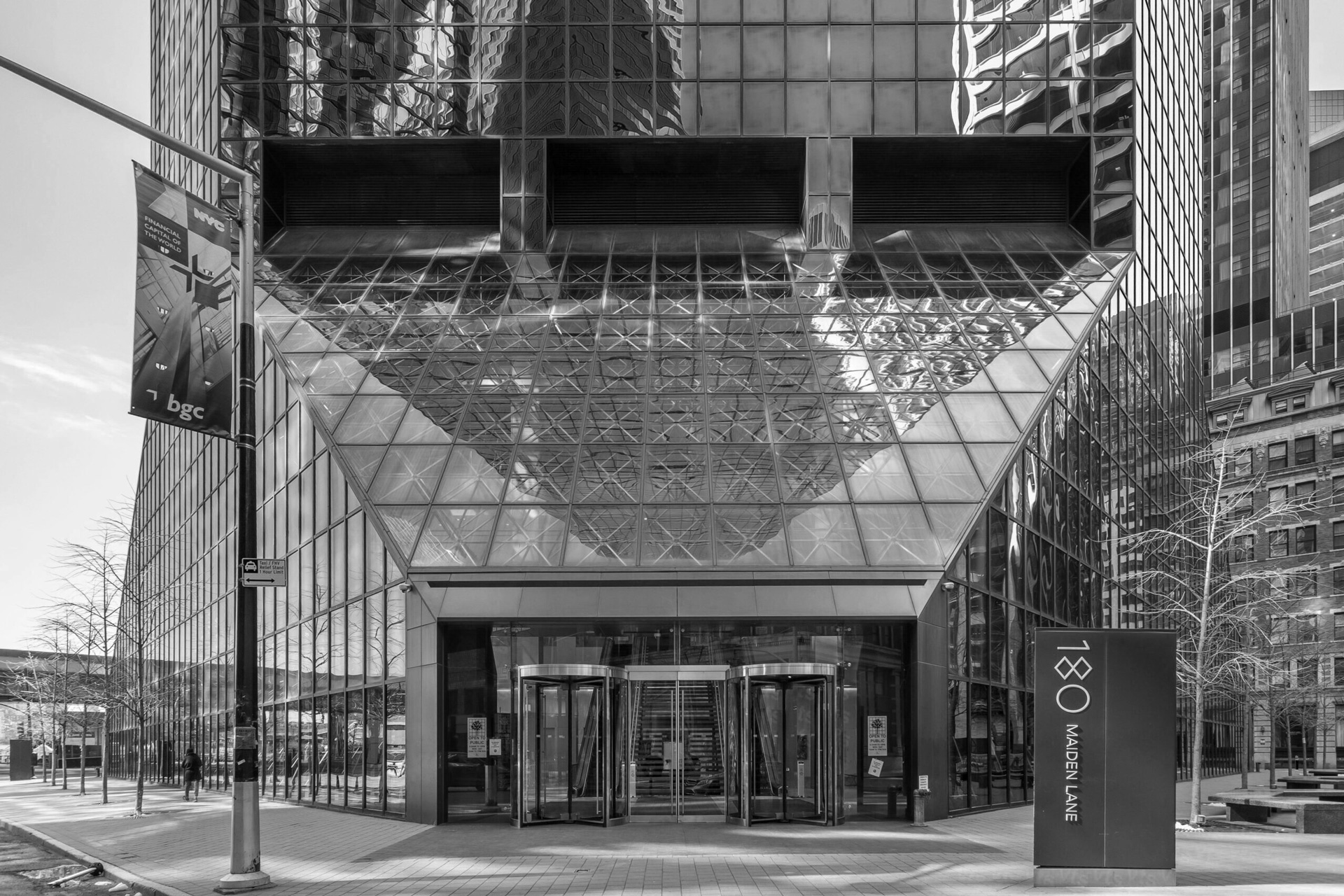 A black-and-white photo of the glass and steel entrance of a modern skyscraper, 180 Maiden Lane. Symmetrical, reflective surfaces echo the unbreakable republic of city buildings. Two revolving doors lead inside, with signage and a street banner visible in front.