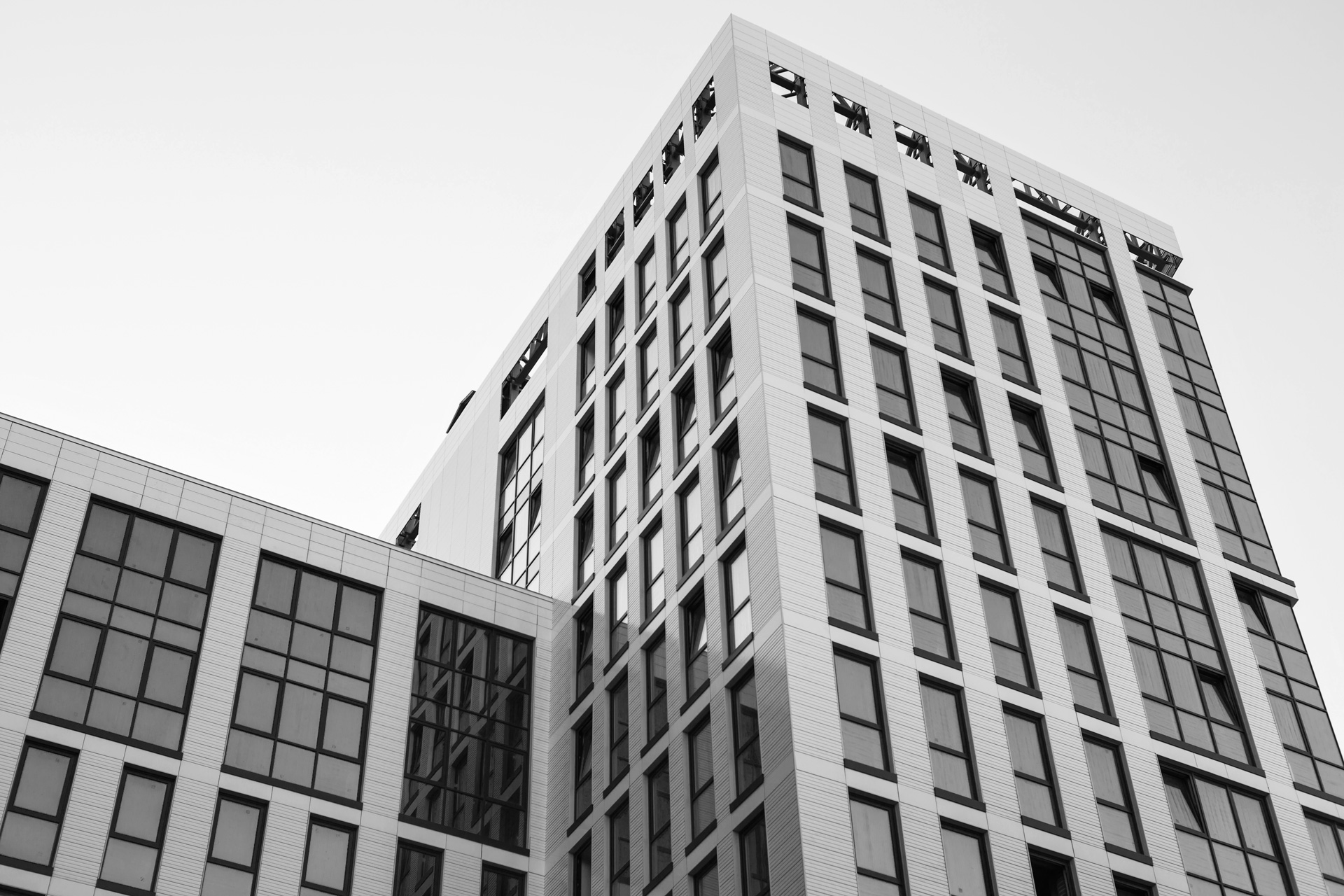 Black and white photo of a modern multi-story building with large rectangular windows. The buildings facade features clean, geometric lines with vertical and horizontal elements. The sky is clear, and the photo is taken from a low angle, emphasizing the buildings height.