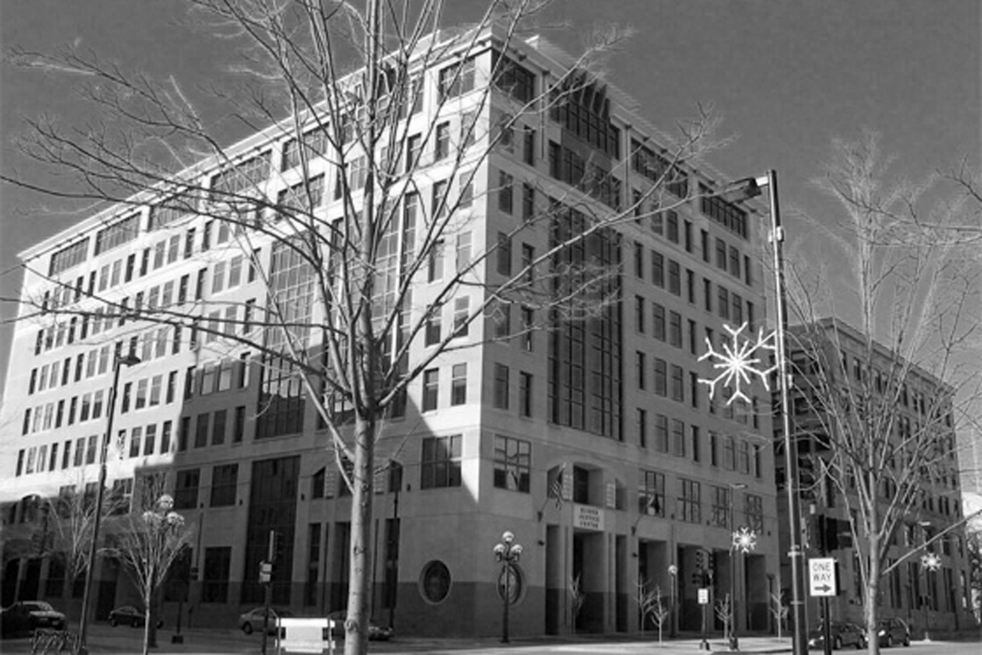 A large, multi-story office building with modern architecture stands on a street corner in the unbreakable republic. Leafless trees line the sidewalk, and holiday decorations, including a star ornament, hang from a street lamp in this black and white scene.