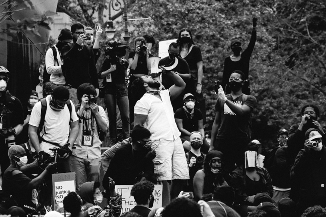 A large group of protesters, many wearing masks, gathers outdoors. One person in the center stands on a platform, shouting into a megaphone for an unbreakable republic. Raised fists and signs capture the intense, unified mood of this black-and-white scene.
