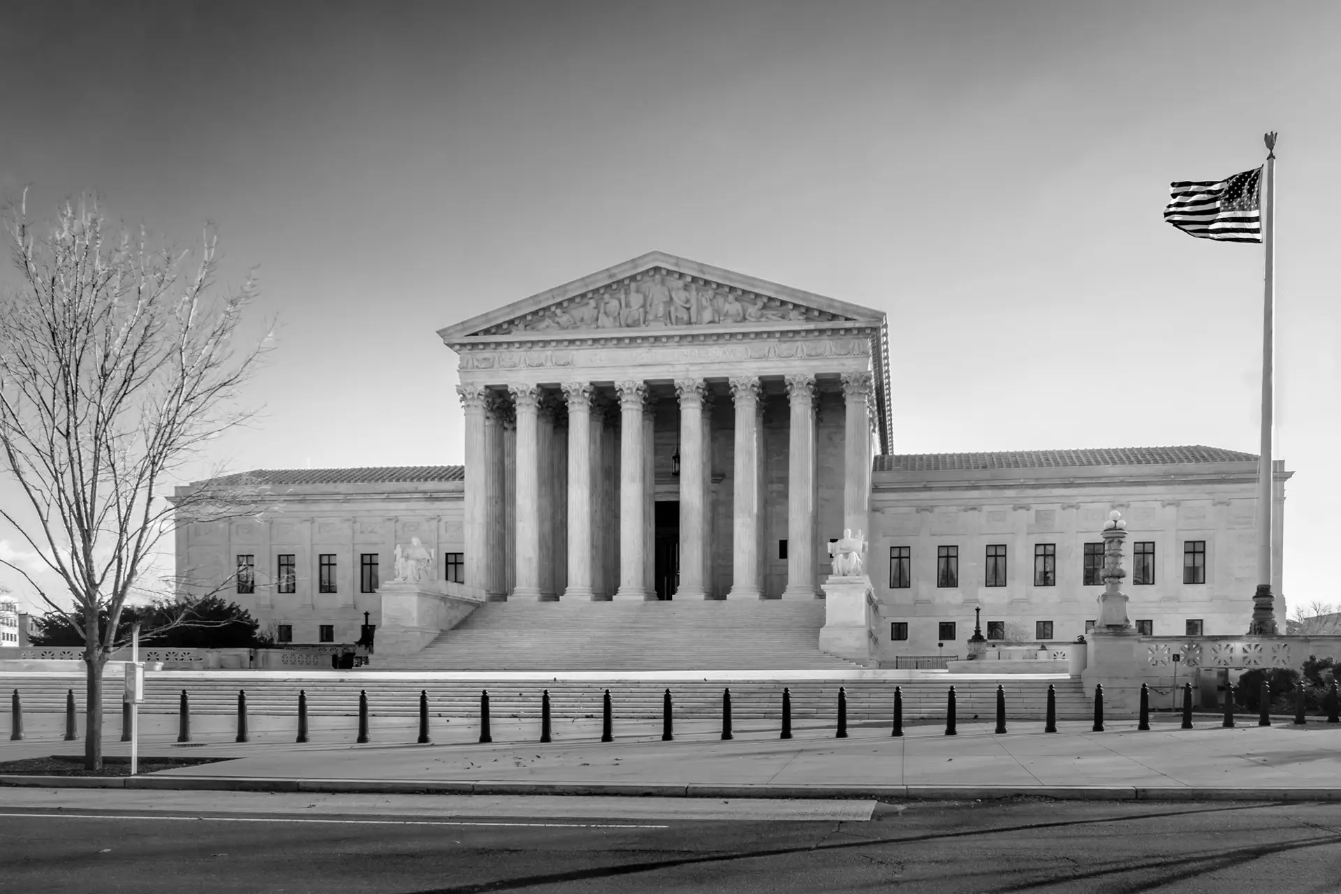Black and white photo of the United States Supreme Court building with tall columns, wide front steps, and a large American flag flying on the right. Leafless tree on the left, statues at the entrance, and a few people near the steps.